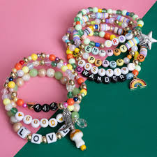 Colorful beaded bracelets on a pink and green background. Each bracelet features words like "LOVE," "PEACE," and "POSITIVITY," with charms such as stars and rainbows.