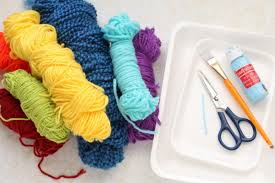 Colorful skeins of yarn in red, yellow, blue, turquoise, and purple are next to a tray holding scissors, a brush, and a paint bottle, evoking creativity.